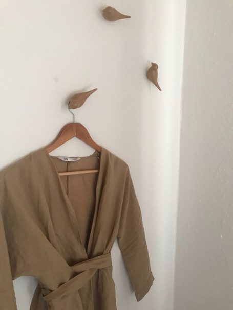 Beige Bademantel auf einem Holzkleiderbügel, dekoriert mit Vogelmotiven an der Wand.